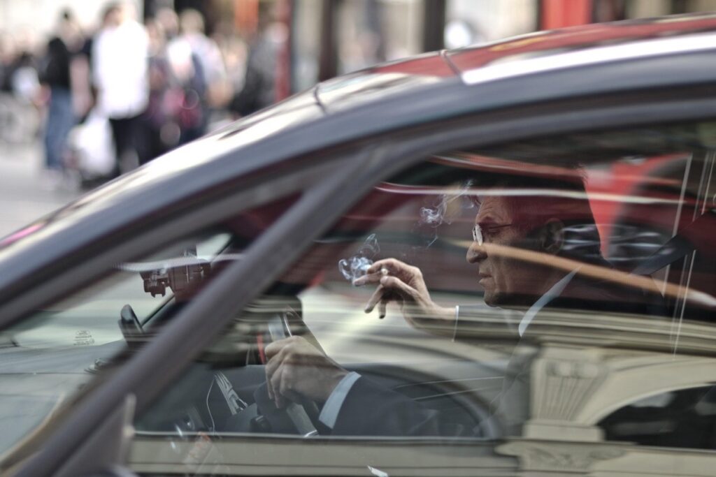 A man in a suit smoking a cigarette while sitting behind the wheel of a car