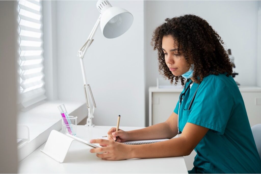 A nurse in scrubs writing notes at a desk in a clinical setting