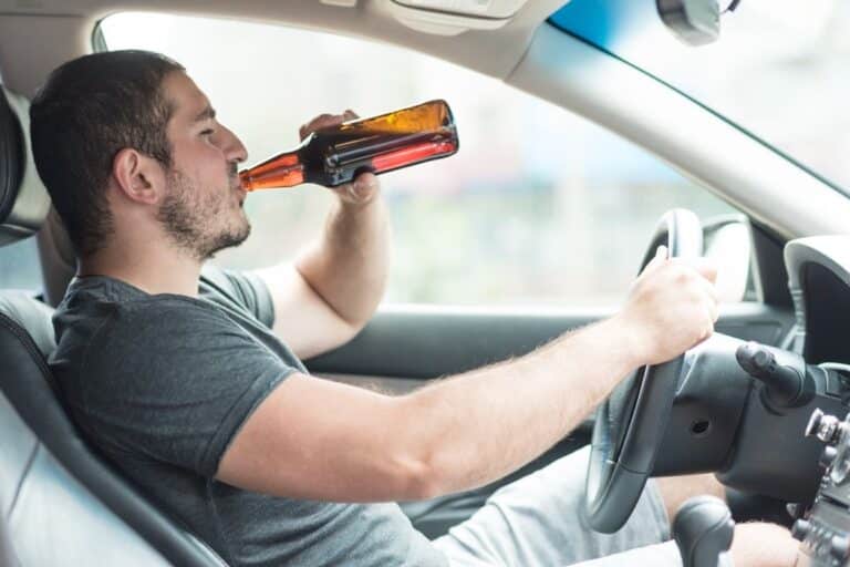 Man drinking from a beer bottle while sitting behind the steering wheel of a car
