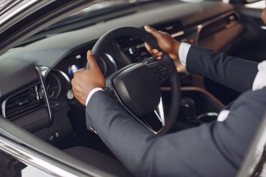 Person in a dark suit sitting behind the steering wheel of a car with both hands on the wheel