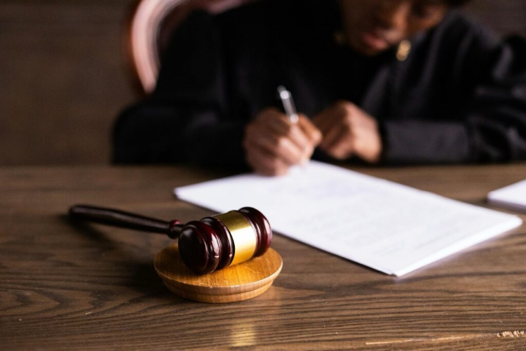 A judge in black robes writes on legal documents beside a wooden gavel on a courtroom bench