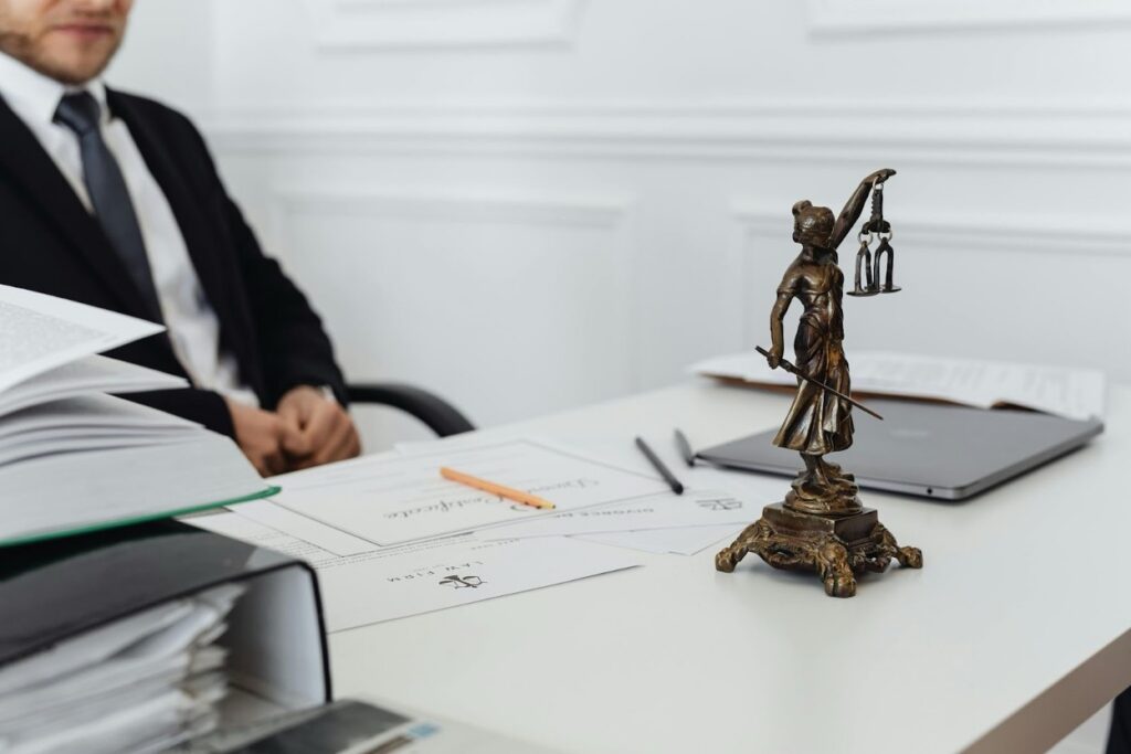 A lawyer seated at a desk with legal documents, a Lady Justice statue, and a laptop.