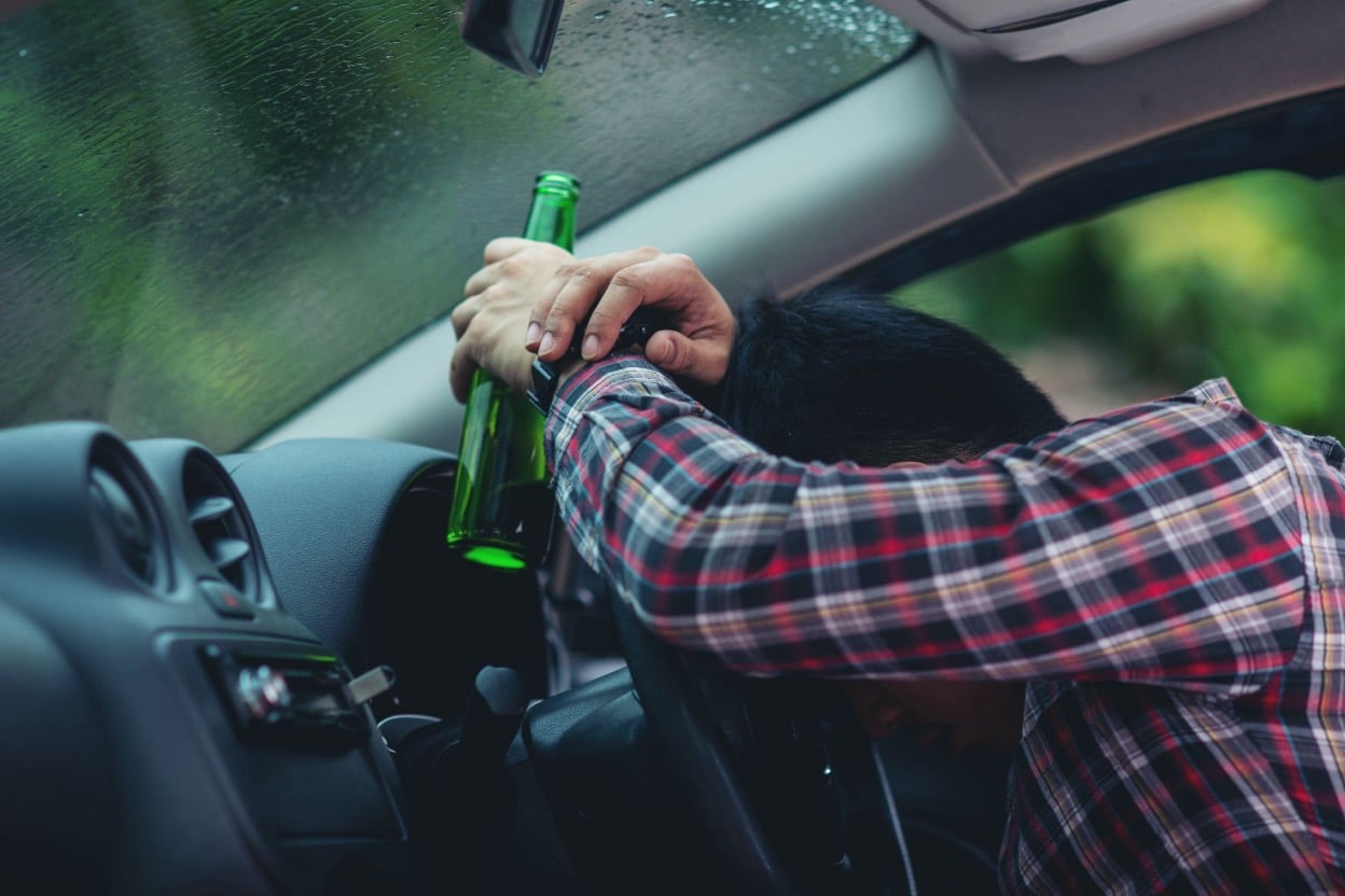 A man slumped over a car's steering wheel while holding a green beer bottle, depicting drunk driving