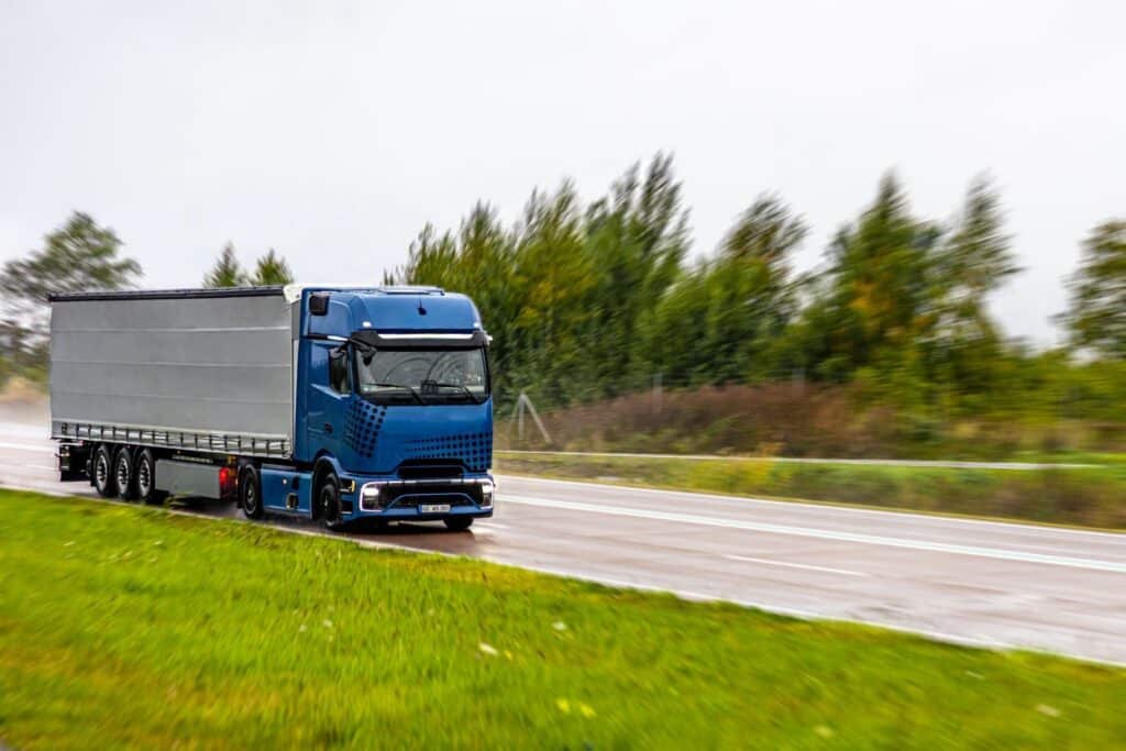 A modern blue semi-truck with a grey curtain-side trailer speeds along a wet highway