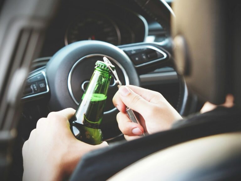 A person sitting in the driver's seat opens a beer bottle with a bottle opener while behind the wheel