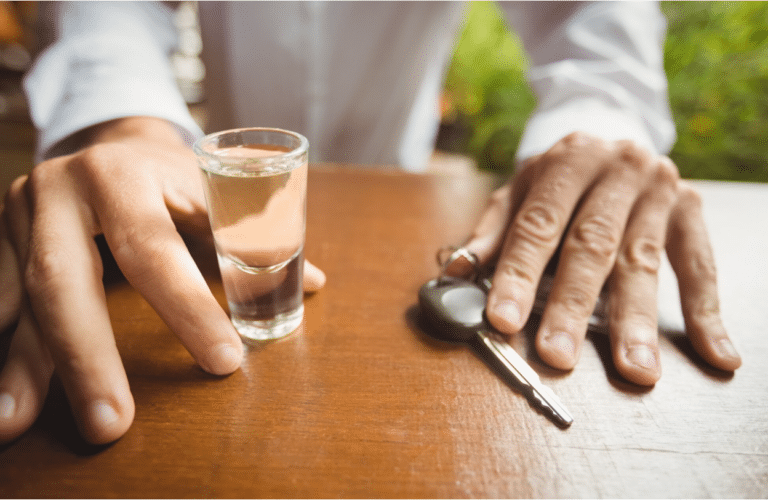 A person's hands resting on a wooden table, one holding a shot glass and the other on car keys