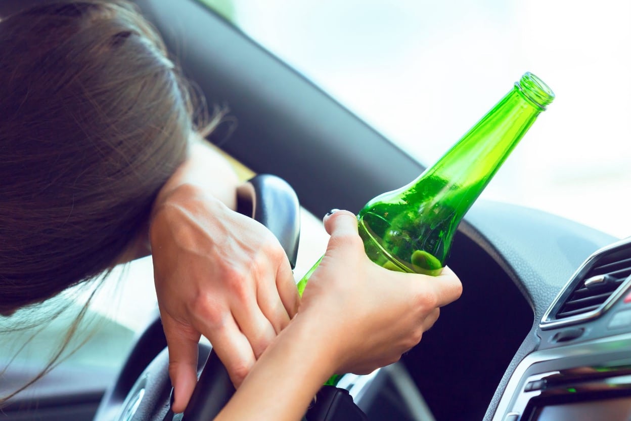 A woman slumped over the steering wheel, holding an open beer bottle while sitting in the driver's seat.