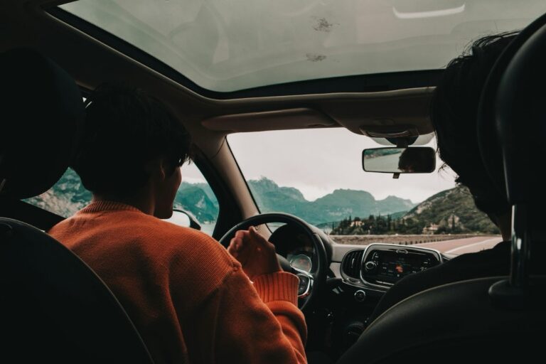 Interior view of a car, showing two people driving toward a scenic mountain range and lake